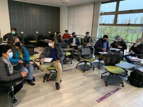 Un grupo de personas con mascarillas se encuentra en un aula moderna, sentados en sillas con ruedas, discutiendo y tomando notas.