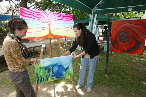 Dos mujeres están en un evento al aire libre, una de ellas sostiene un lienzo mientras la otra lo observa entre pinturas coloridas en un entorno natural.