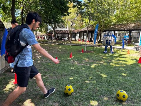 Un joven patea un balón de fútbol en un área al aire libre con decoraciones y un personaje de una mascota en el fondo.