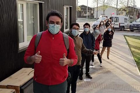 Un grupo de personas en fila, usando mascarillas, mientras esperan en un lugar exterior.