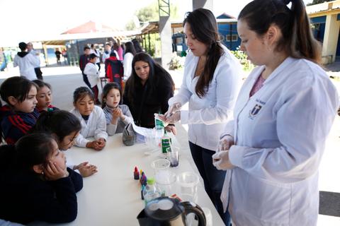 Un grupo de niños observa atentamente a dos mujeres que realizan un experimento científico al aire libre.