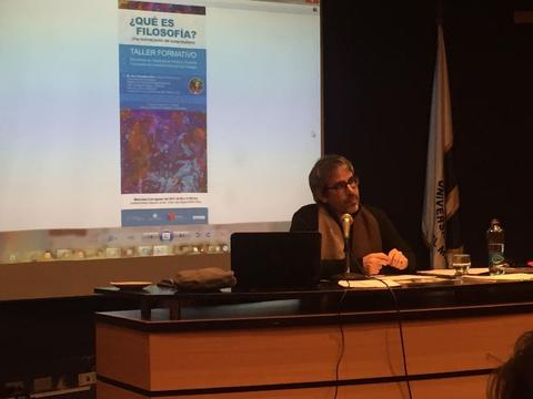 Un hombre hablando en una conferencia sobre filosofía frente a una pantalla que muestra un cartel del taller formativo.