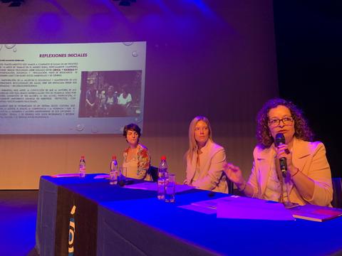 Tres mujeres participan en una mesa redonda mientras una presenta diapositivas en el fondo.