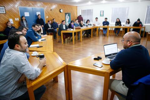 Un grupo de personas asiste a una reunión en un salón con mesas de madera dispuestas en forma de U.