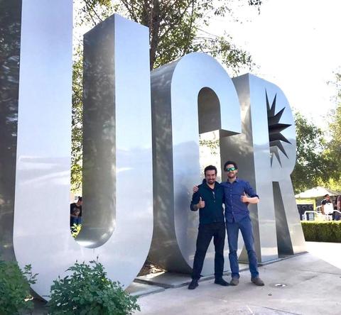Dos hombres sonrientes posan junto a un gran letrero metálico que dice 'UCR'.