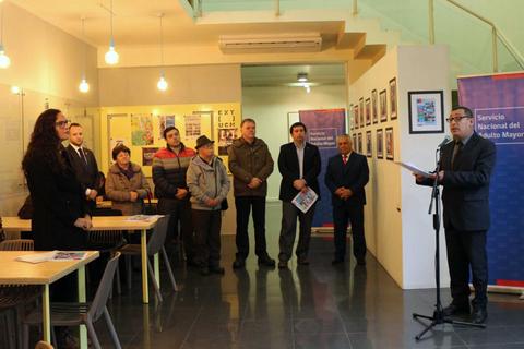 Un evento inaugurado con varias personas escuchando a un orador en un salón decorado con fotografías.