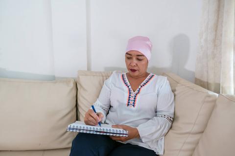 Una mujer con pañuelo en la cabeza escribe en un cuaderno mientras está sentada en un sofá claro.