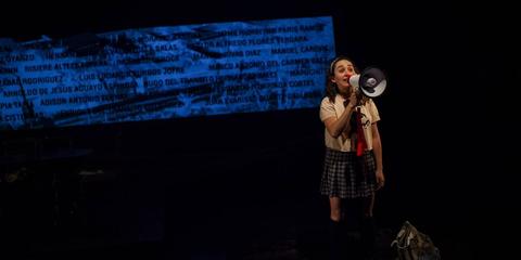 Una joven con un megáfono se presenta en un escenario oscuro, mientras una proyección de texto se muestra de fondo.