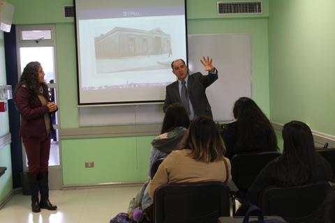 Un hombre está dando una presentación en un aula, mientras una mujer lo escucha atentamente.
