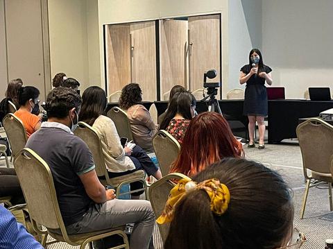 Una presentadora habla frente a un grupo de personas en un evento en una sala de conferencia.