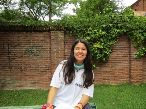 Una joven sonriente sentada en un banco de un parque, rodeada de vegetación y un muro de ladrillos.