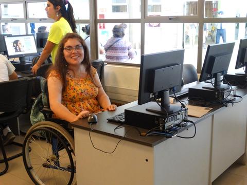 Una mujer sonriente en silla de ruedas trabaja en una computadora en un aula.