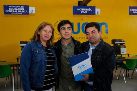 Un grupo de tres personas sonríe delante de un cartel de la Universidad Católica del Maule.