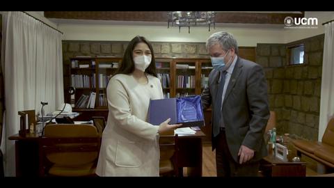 Una mujer y un hombre, ambos con mascarillas, están en una oficina, donde la mujer presenta un objeto en una caja azul al hombre.