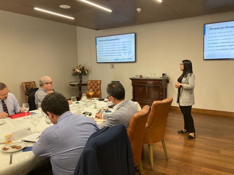 Una mujer está presentando información en una reunión mientras varias personas la escuchan atentamente alrededor de una mesa.