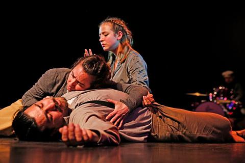 Un grupo de tres personas en una escena dramática, con una mujer abrazando a un hombre tendido en el suelo mientras otra mujer observa.