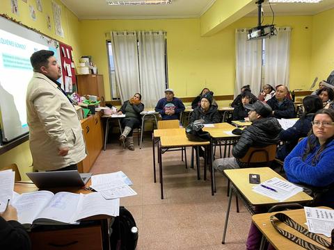 Un grupo de personas se encuentra en un aula escuchando a un presentador que está de pie frente a una pantalla.