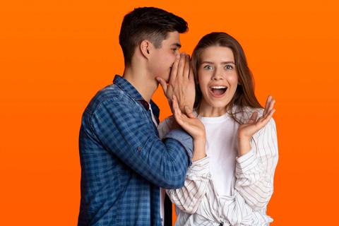 Un hombre susurra algo a una mujer que sonríe emocionada frente a un fondo naranja.