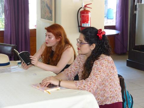 Dos mujeres están sentadas en una mesa, una lee un libro mientras la otra observa.