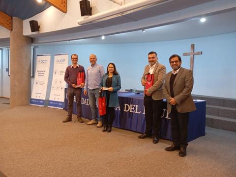 Un grupo de cinco personas posando en un evento en un salón con un atril y banners de la universidad al fondo.