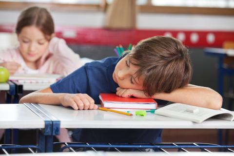 Un niño dormido en su escritorio durante una clase, mientras una niña estudia al fondo.