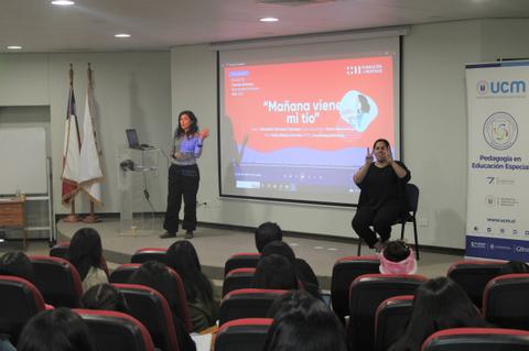 Una presentación educativa con una ponente y una intérprete en un auditorio lleno de estudiantes.