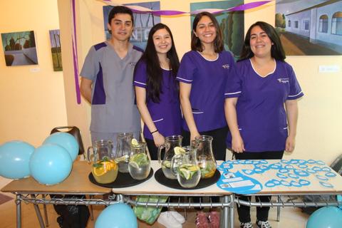 Un grupo de cuatro personas vestidas con uniformes morados sonríe frente a una mesa con bebidas y globos azules en un ambiente festivo.