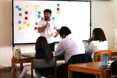 Una persona está presentando frente a un grupo de personas en un entorno de aula con post-its en la pizarra.