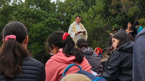 Un grupo de personas escucha a un sacerdote que está leyendo un texto en un entorno natural.