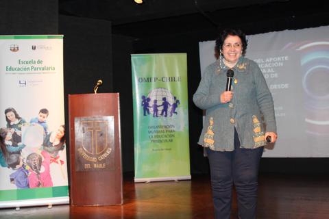 Una presentadora sonriente se encuentra en un escenario con materiales educativos detrás de ella.