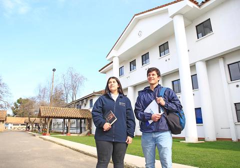 Dos estudiantes posan frente a un edificio educativo en un día despejado.