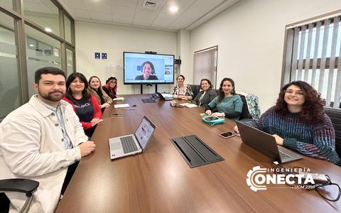 Un grupo de personas se encuentra en una sala de reuniones, algunos usando computadoras portátiles y otros participando a través de videoconferencia.