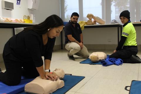 Una mujer está practicando resucitación cardiopulmonar en un maniquí mientras dos hombres observan en un aula.