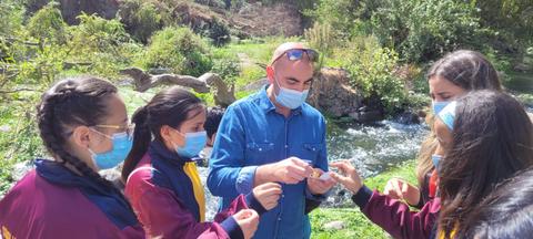 Un grupo de personas con mascarillas se encuentra en un entorno natural, interactuando entre sí junto a un río.