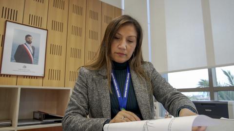 Una mujer revisa documentos en un escritorio con una fotografía de un hombre en la pared de fondo.