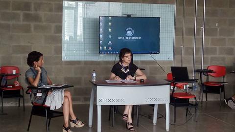 Una reunión en un salón moderno con dos mujeres sentadas en una mesa frente a una pantalla mostrando el texto 'LOS LIBERADORES'.