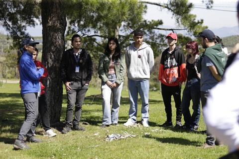Un grupo de jóvenes escucha atentamente a un orador bajo un árbol en un entorno natural.