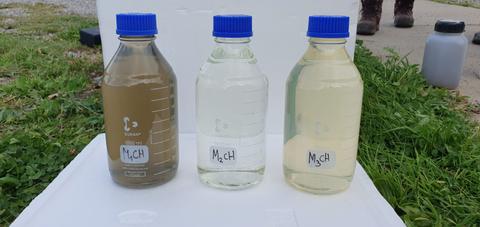 Tres botellas de líquido con diferentes colores sobre una superficie blanca y césped.