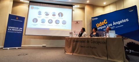 Una presentación en un aula con un proyector mostrando información y tres personas sentadas en una mesa.