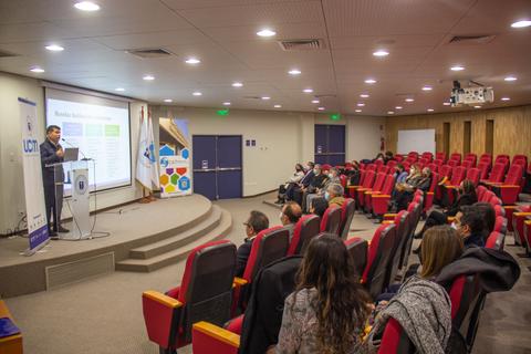 Una conferencia en un auditorio con un ponente frente a una audiencia sentada en sillas rojas.