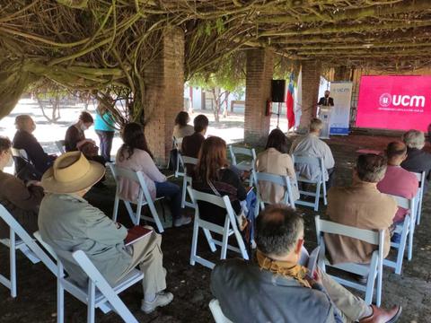 Una reunión en un espacio al aire libre con varias personas sentadas frente a un escenario donde se presenta un logo de UCM.