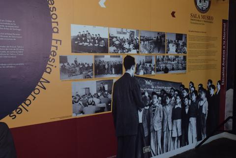 La imagen presenta una exposición con fotografías históricas en una sala de museo dedicada a la maestra Fresia Morales.