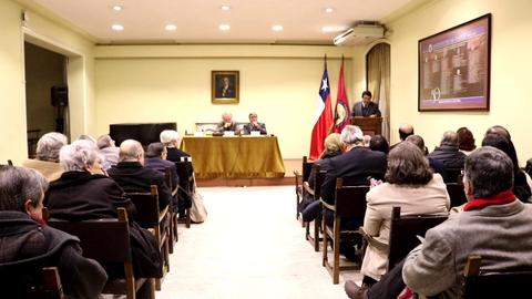 Una reunión formal con una audiencia sentada, un ponente en el estrado y banderas chilenas presentes.