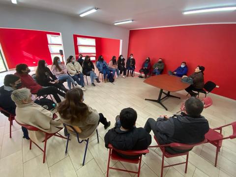 Un grupo de personas sentadas en círculo en una sala con paredes rojas y ventanas, participando en una reunión.
