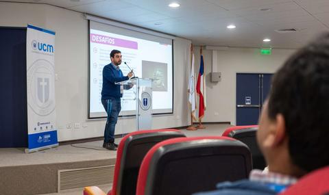 Un presentador expone en un auditorio frente a una audiencia mientras se proyecta una presentación en la pantalla.