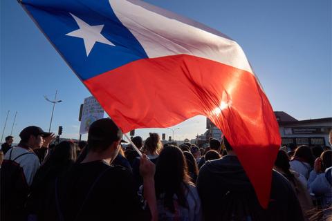 Una multitud sostiene una bandera de Chile mientras se manifiestan bajo un cielo despejado.