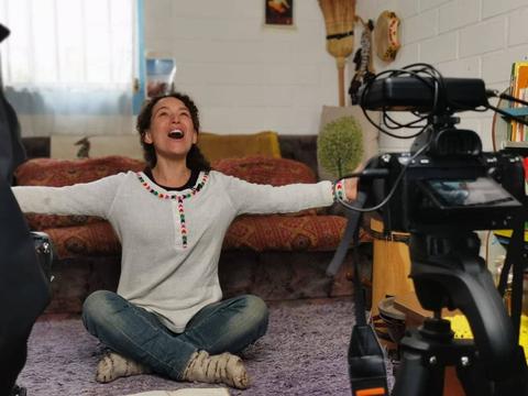 Una mujer sonriente se sienta en el suelo con los brazos extendidos en un ambiente acogedor.