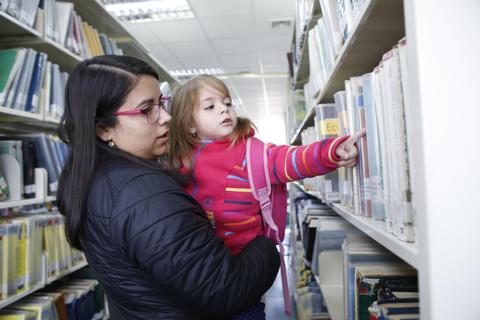 Una madre sostiene a su hija en una biblioteca mientras la niña señala un libro en las estanterías.