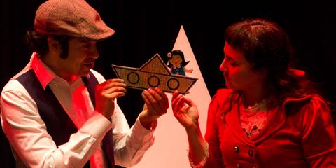Dos personas sostienen un barco de cartón mientras interactúan entre sí en un ambiente teatral.