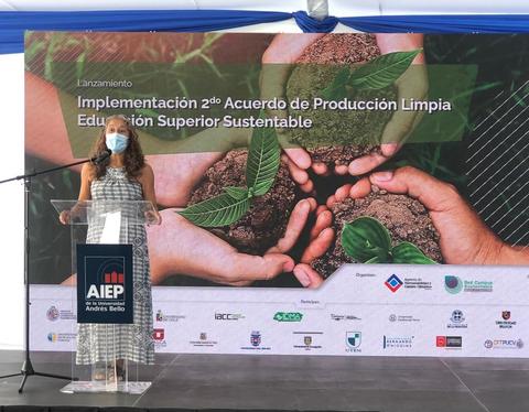 Una mujer habla en un evento sobre producción limpia y educación sustentable frente a un fondo gráfico de manos con plantas.